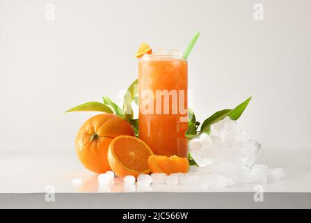 Orange slushie in tall glass with fruit and crushed ice around it on white table with isolated background. Front view. Horizontal composition. Stock Photo