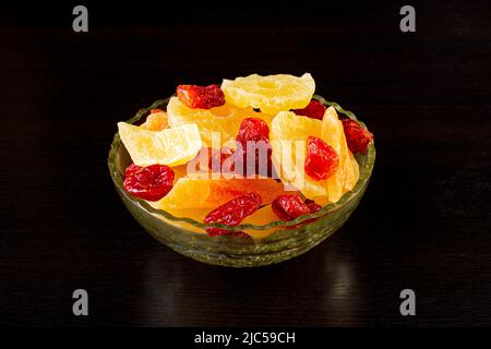 Yellow candied melon slices, pineapple rings and dogwood in a glass vase on a black background Stock Photo