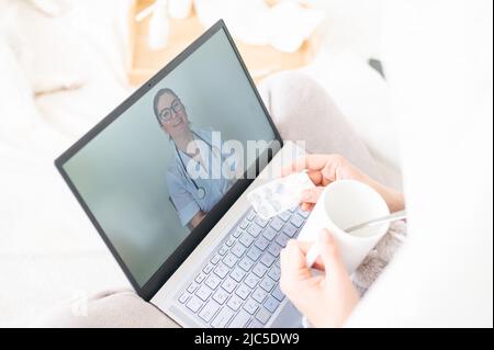 A woman lies in bed and holds a spray for sore throat. A friendly doctor talks to a sick patient by video link. Therapist on a laptop screen. Online Stock Photo