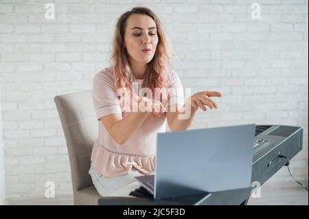 A friendly woman plays the electronic piano and conducts a video blog on her laptop. Stay home. Musical instrument teacher. Distance learning music Stock Photo