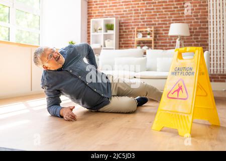 Mature Man Falling On Wet Floor In Front Of Caution Sign At Home Stock Photo
