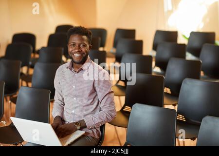 Portrait of cheerful handsome young black manager in shirt sitting on chair in conference room and using laptop Stock Photo