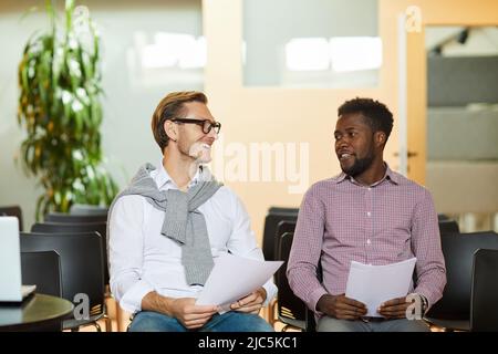 Smiling young multi-ethnic specialists sitting on chairs in auditorium and sharing ideas of marketing strategy Stock Photo