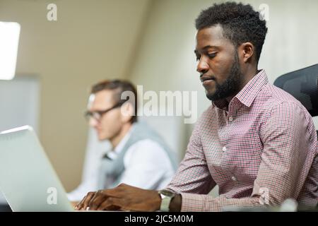 Concentrated young African-American programmer in checkered shirt sitting at table and typing on laptop Stock Photo