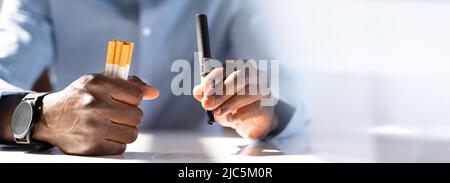 Man Holding Vape And Tobacco Cigarette Over Desk Stock Photo