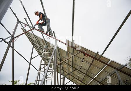 Man engineer standing on metal rail and using electric drill while installing support structure for photovoltaic solar panels. Isolated on white background. Stock Photo