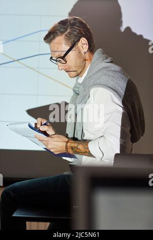Content experienced financial coach in smart casual outfit sitting on table against projection screen and making notes in paper Stock Photo