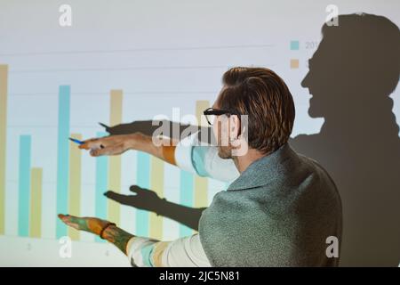 Rear view of modern businessman with earring pointing at graph on projection screen while explaining sales strategy Stock Photo