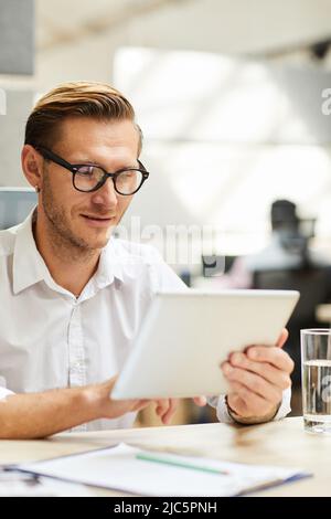Content clever young businessman in eyeglasses sitting at table and using tablet browsing internet Stock Photo