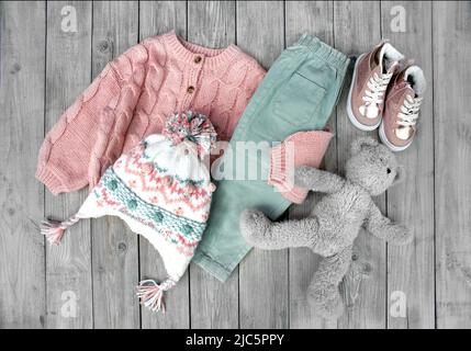 Child's girl knitwear set on wooden background flat lay. Kid's winter woolen clothes top view. Toddler knitted cardigan,pants,hat and shoes. Collectio Stock Photo