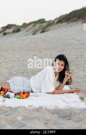 Young woman holding wine and looking at camera near sunglasses and fruits on beach Stock Photo