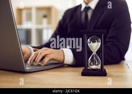 Close up of black hourglass with white trickling sand on table of businessman working on laptop. Stock Photo