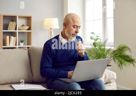 Senior man sitting on couch at home, working on his laptop computer and thinking Stock Photo