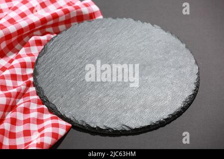 Slate black round dish empty space on dark background decorated with checkered cloth.Tableware,sushi plate.Top view backdrop. Stock Photo