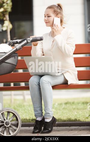 Young mother in white coat working outside on her smartphone and laptop while baby sleeping in stroller Stock Photo