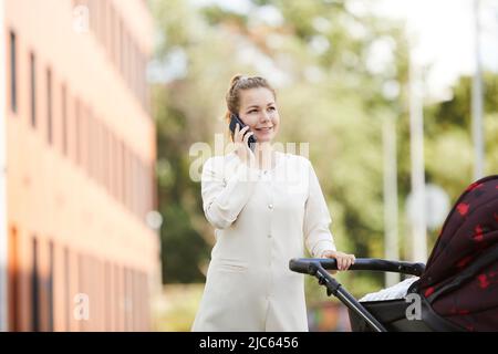 Young mother in white coat calling her friends while walking with her baby who is playing in stroller Stock Photo
