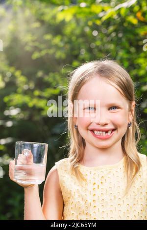 Happy little girl wearing yellow dress holding glass cup of water in a summer garden - healthy lifestyle concept Stock Photo