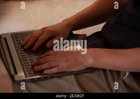 a man's hands on a computer keyboard. a white-skinned man types on the keys of a laptop keyboard. He is sitting on a white sofa. We can't see his face Stock Photo