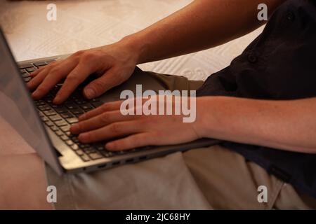a man's hands on a computer keyboard. a white-skinned man types on the keys of a laptop keyboard. He is sitting on a white sofa. We can't see his face Stock Photo