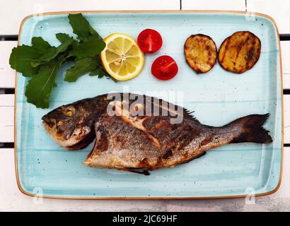 Fried fish with vegetables and herbs lies on light blue plate - tray, top view, close-up. Restaurant decoration of fish serving. Stock Photo