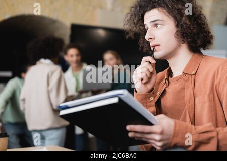 thoughtful student with copybooks looking away near classmates in blurred auditorium Stock Photo