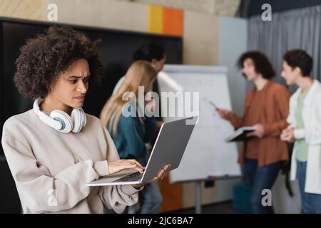 tense african american woman using laptop near multiethnic students talking on blurred background Stock Photo