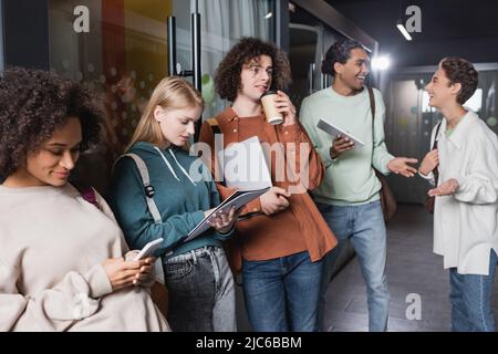 young man with laptop drinking coffee near interracial friends in hallway of university Stock Photo