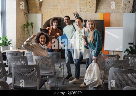 excited multiethnic classmates shouting and showing success gesture in auditorium Stock Photo