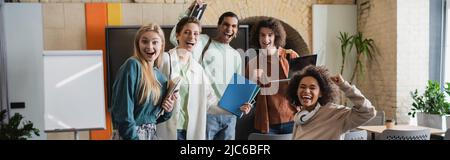 excited interracial students with copybooks showing win gesture in classroom, banner Stock Photo