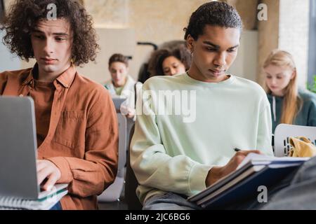 african american student writing in notebook while friend typing on laptop during lesson Stock Photo