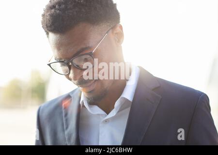 Close-up of content handsome young African businessman in glasses wearing suit looking down outdoors Stock Photo