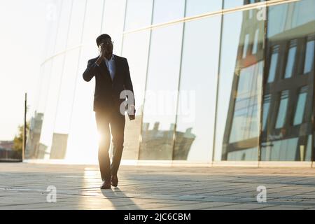 Contemporary mobile black manager in suit walking over financial district and talking to client by phone Stock Photo