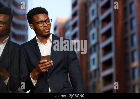 Serious young Afro-American businessman with hipster beard leaning on building wall and texting message on smartphone Stock Photo