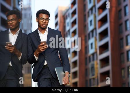 Serious confident African-American businessman in eyeglasses standing on city street and checking notes in smartphone Stock Photo