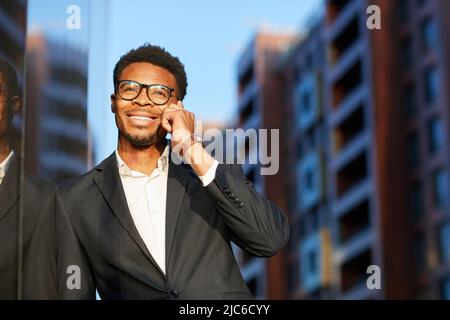 Cheerful young Black businessman in suit standing by contemporary building and chatting on phone Stock Photo