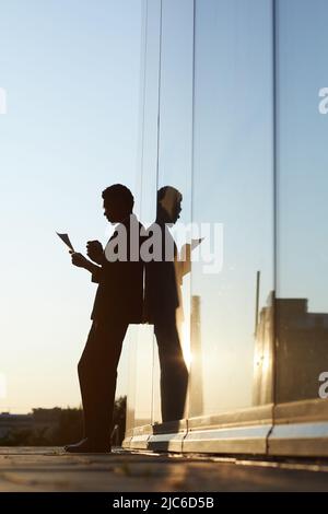 Silhouette of young businessman busy with papers leaning on building outdoors and drinking coffee while examining papers Stock Photo