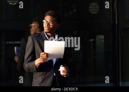 Serious pensive young Afro-American man with beard standing by office building and drinking coffee while reading papers Stock Photo