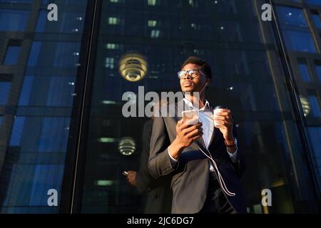 Low angle view of serious thoughtful young African-American businessman in eyeglasses standing by city building and using smartphone in earphones Stock Photo