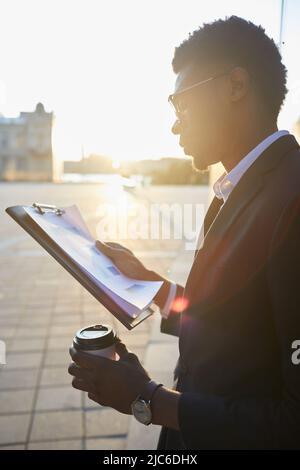 Serious young Afro-American businessman in eyeglasses walking over street and drinking coffee while analyzing sales graphs on move Stock Photo