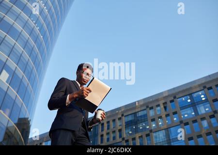 Below view of thoughtful young black businessman in elegant suit walking over business district and examining papers on move Stock Photo