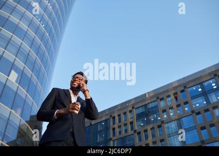 Smiling contemporary young black businessman in suit talking by mobile phone and drinking coffee while walking over street Stock Photo