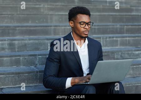 Serious concentrated young African-American man in suit sitting on stairs and typing on laptop while working on report out of office Stock Photo