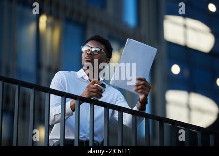 Serious thoughtful young black businessman in white shirt leaning on railing and drinking coffee while analyzing documents in dark lobby Stock Photo