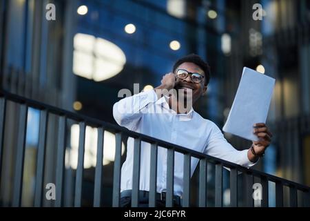 Cheerful excited young black businessman in glasses standing on balcony of business center and holding papers while talking by phone Stock Photo