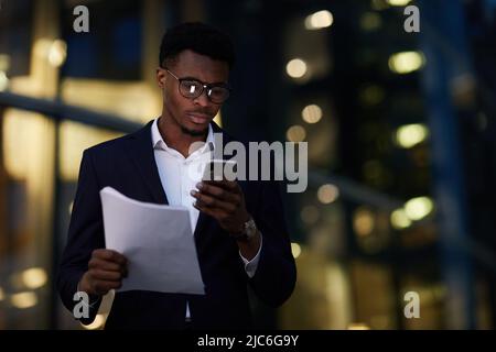 Serious busy young black man in eyeglasses standing against office building at night and holding papers while checking information on internet Stock Photo