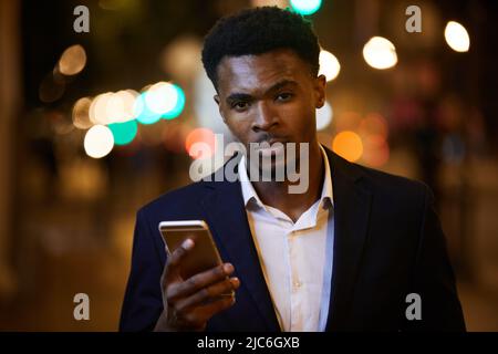 Portrait of serious handsome young black businessman with beard wearing suit and white shirt checking phone while walking over night city Stock Photo