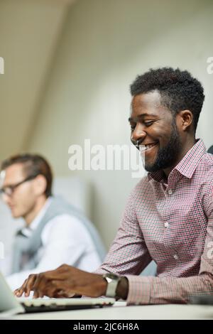 Cheerful handsome young Black programmer satisfied with web project sitting at desk in office and typing on laptop Stock Photo
