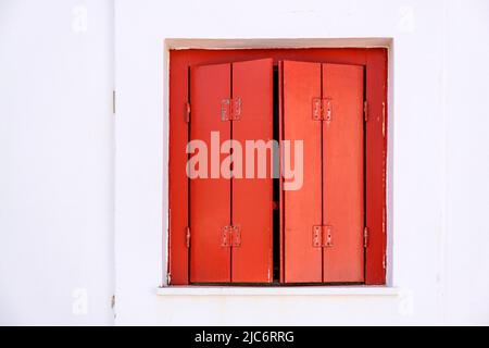 Window with semi closed wooden shutters red color on white wall background. Cyclades island house front view, Greek traditional architecture. Stock Photo