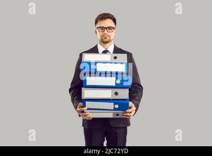 Serious male office worker standing on gray background holding stack of large folders for documents. Stock Photo