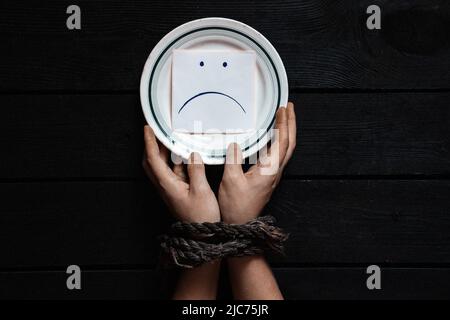 a plate with paper on which is drawn a sad smiley and women's hands tied with a rope, hunger and nothing to eat, slavery Stock Photo